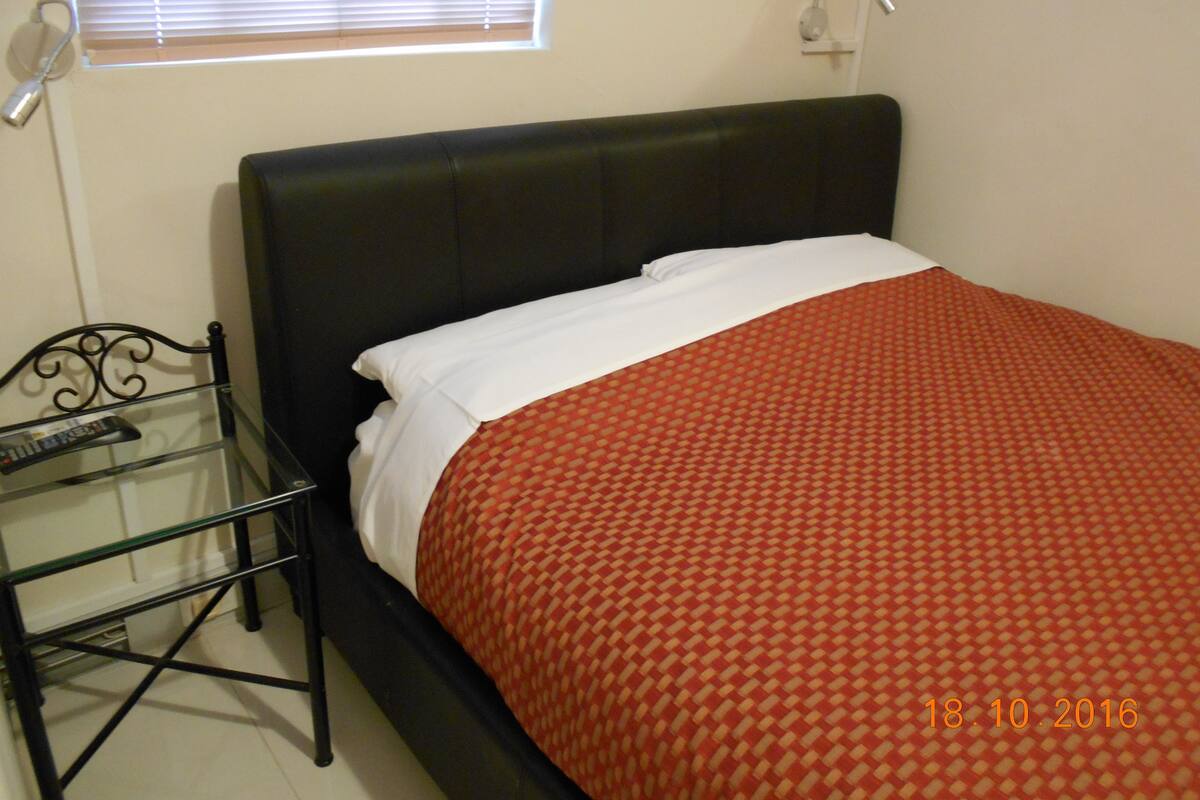 Une chambre à coucher avec une tête de lit noire, une table de chevet en verre, une couvre-lit rouge à motifs et un oreiller blanc.
