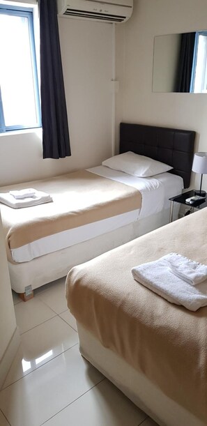 A hotel room with two beds, a window with curtains, a wall-mounted air conditioner, a bedside table with a lamp, and a mirror.