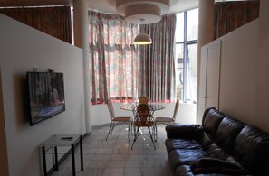 A living room with a leather couch, a TV mounted on the wall, a dining table with chairs, and a window with curtains.