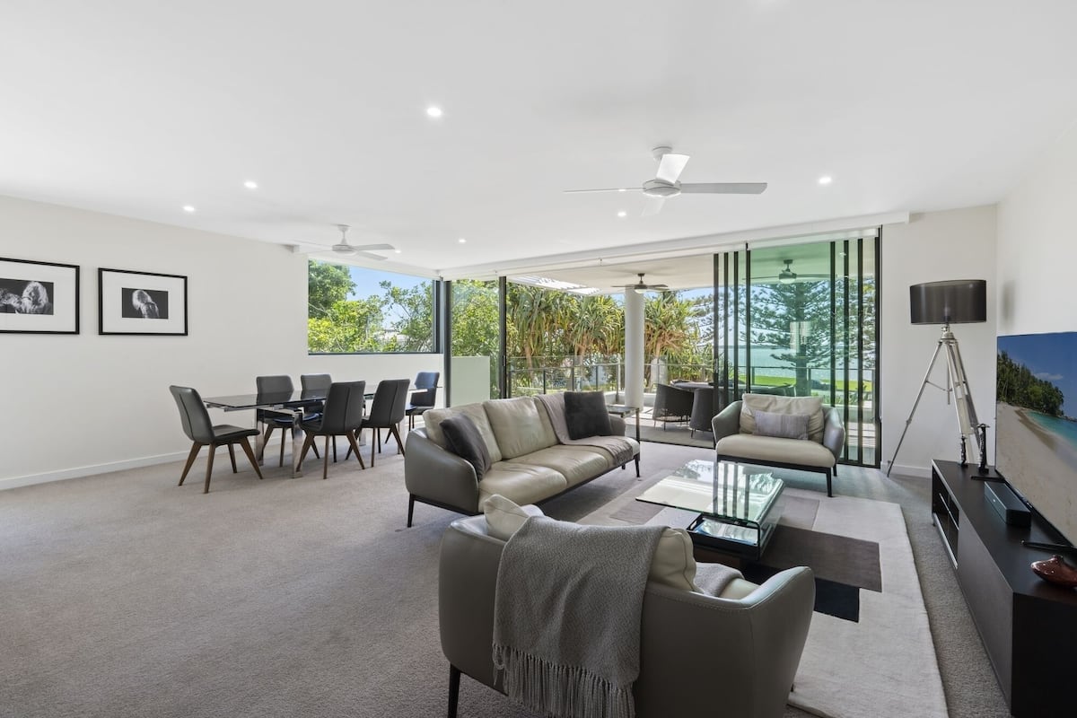 A modern living room with a sofa, coffee table, and dining area. There is a balcony with a view of trees and a beach.