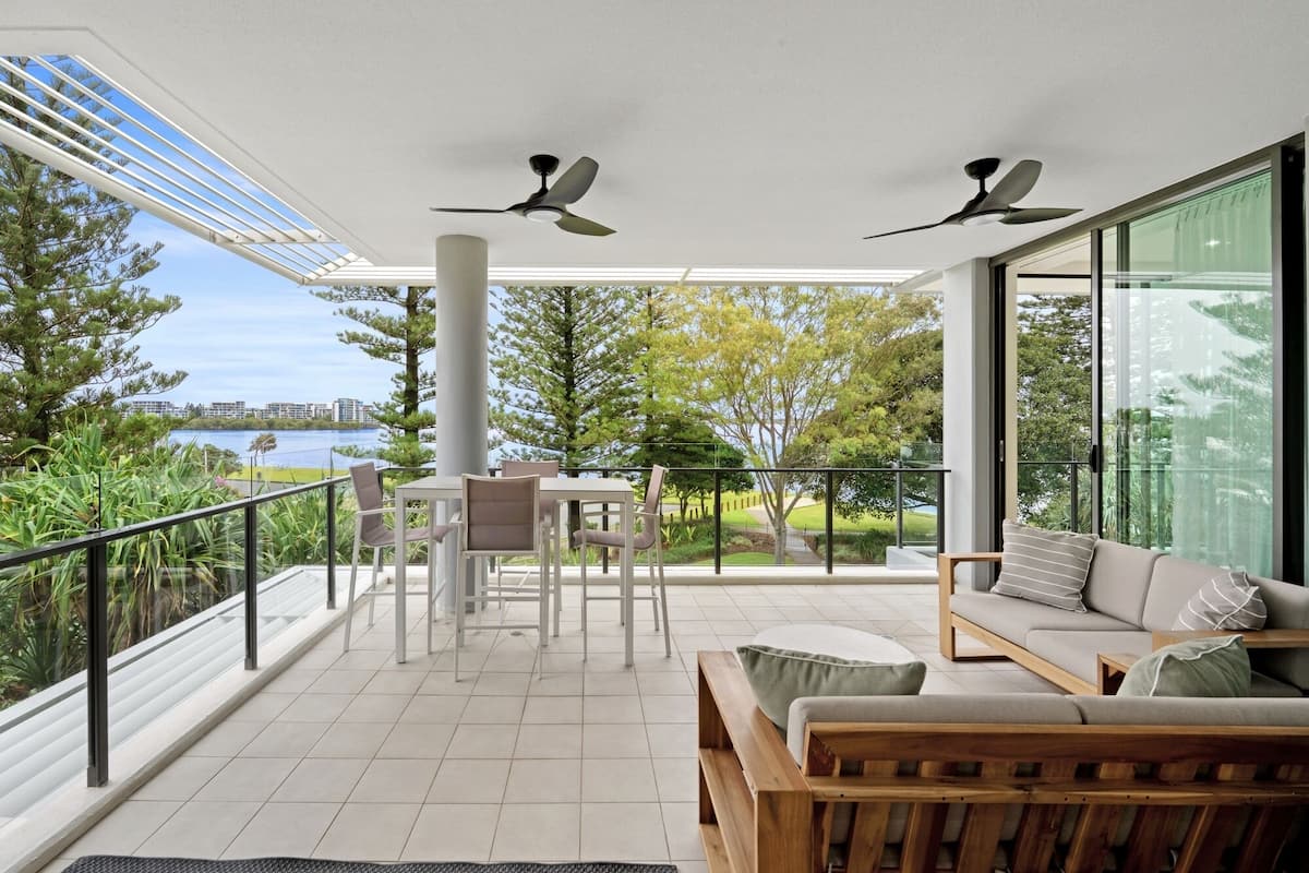 A balcony with a table and chairs, a sofa, two ceiling fans, and a view of a lake and buildings.