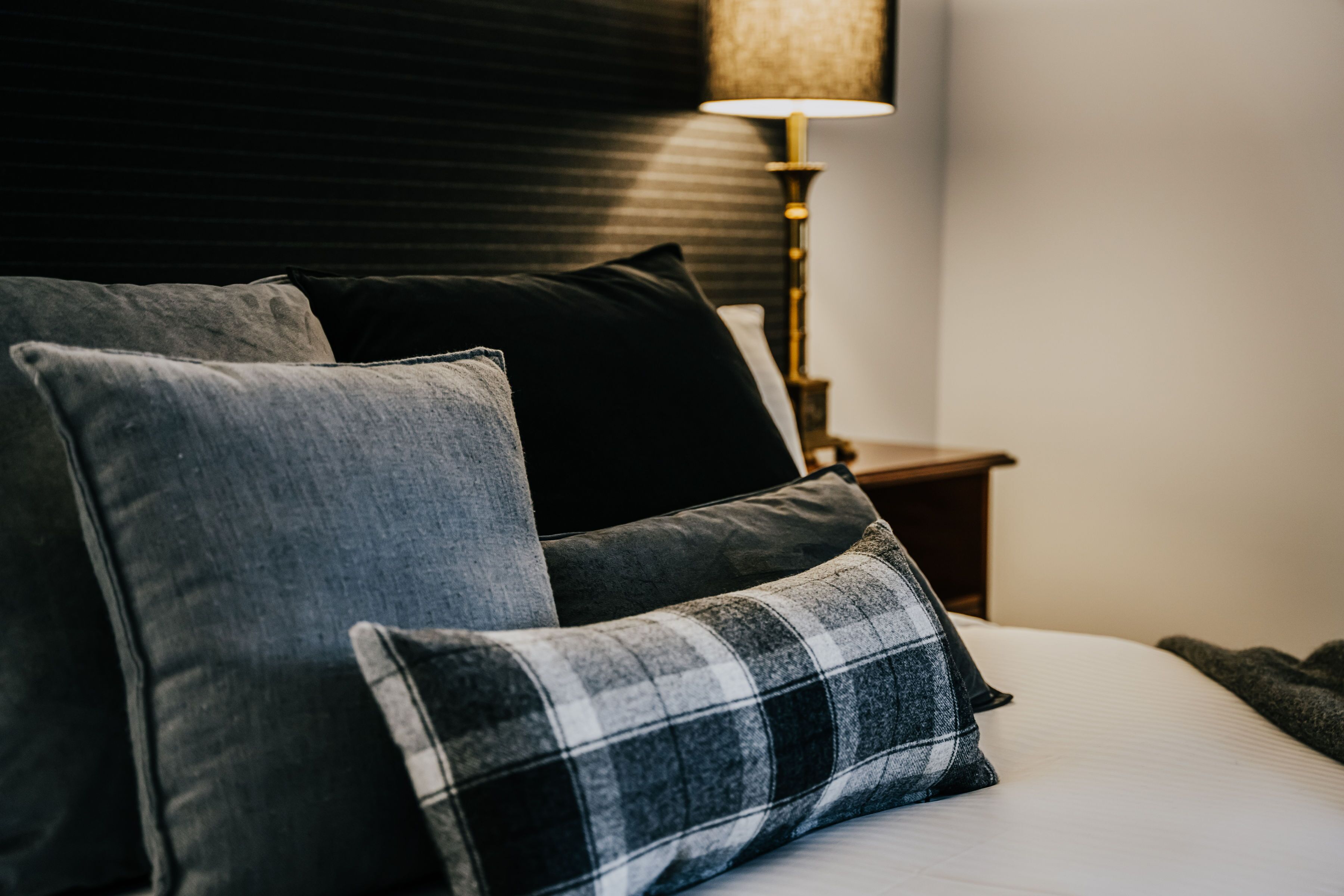 Una cama bien hecha con almohadas grises y negras, una almohada a cuadros y una lámpara de noche sobre una mesita de madera.