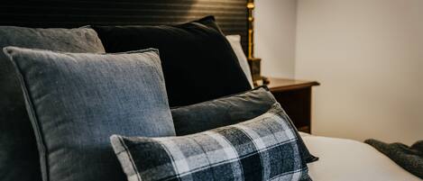 Una cama bien hecha con almohadas grises y negras, una almohada a cuadros y una lámpara de noche sobre una mesita de madera.