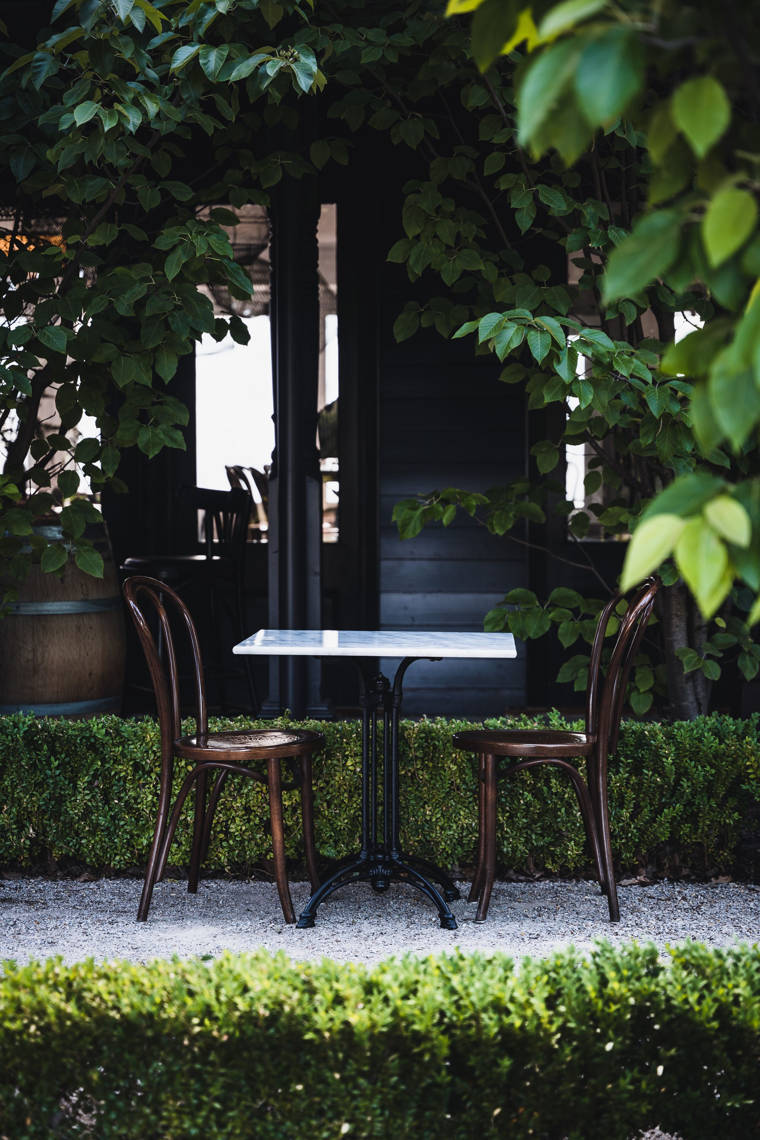An outdoor seating area with a table and two chairs, surrounded by greenery and a building in the background.