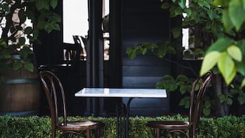 An outdoor seating area with a table and two chairs, surrounded by greenery and a building in the background.