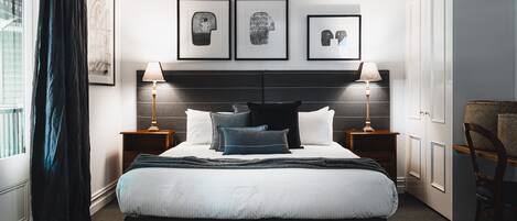 Una habitación de hotel moderna con una cama grande, dos mesitas de noche con lámparas, un cabecero con obra de arte y vistas a la ciudad desde la ventana.