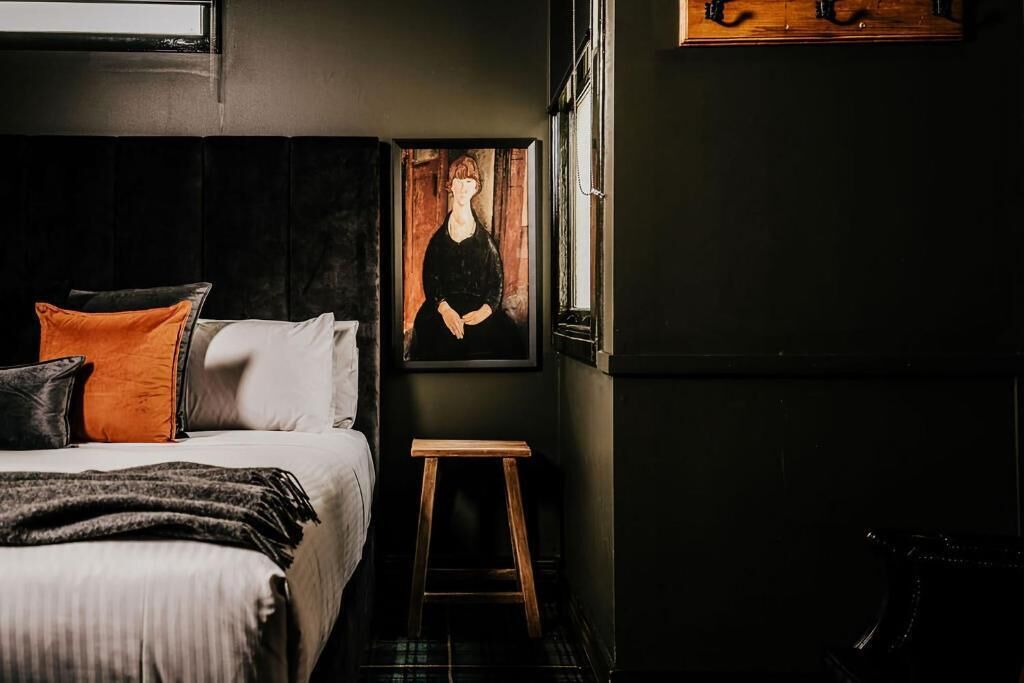 Un dormitorio con cama, almohadas, un taburete de madera y un cuadro en la pared.