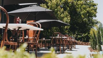Outdoor tables and chairs with umbrellas, a brick wall, and a large tree in the background.
