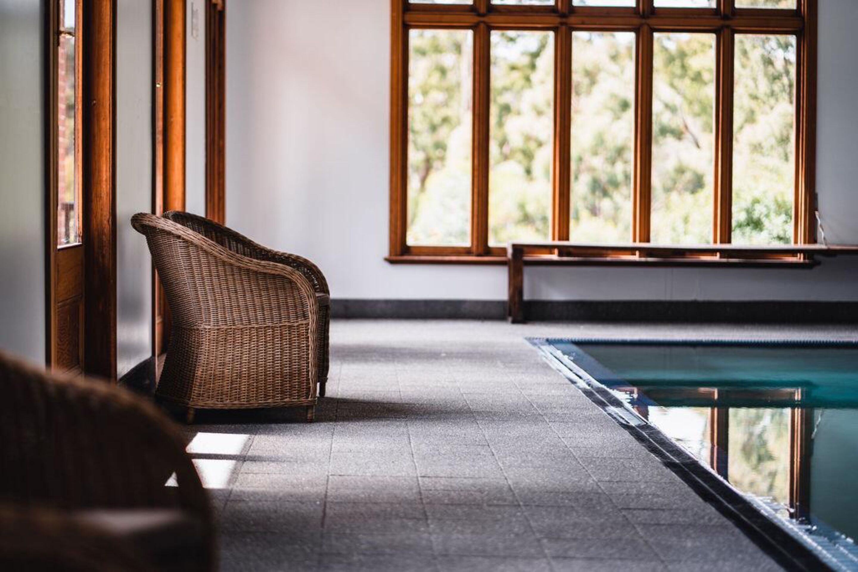 A room with a wicker chair, a pool, and a window with a view of greenery.