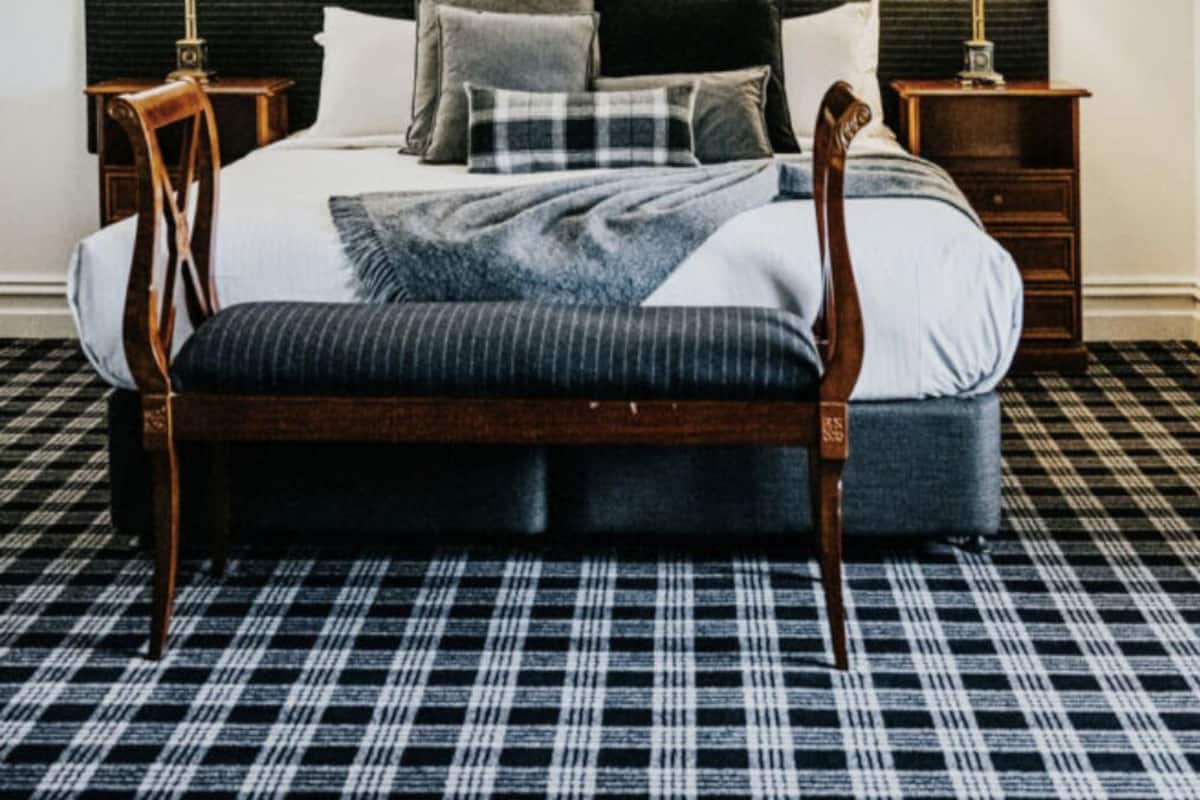 Una cama bien tendida con un cabecero oscuro, flanqueada por dos mesitas de noche con lámparas, un suelo a cuadros y tres cuadros enmarcados sobre la cama.