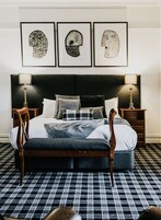 Una cama bien tendida con un cabecero oscuro, flanqueada por dos mesitas de noche con lámparas, un suelo a cuadros y tres cuadros enmarcados sobre la cama.