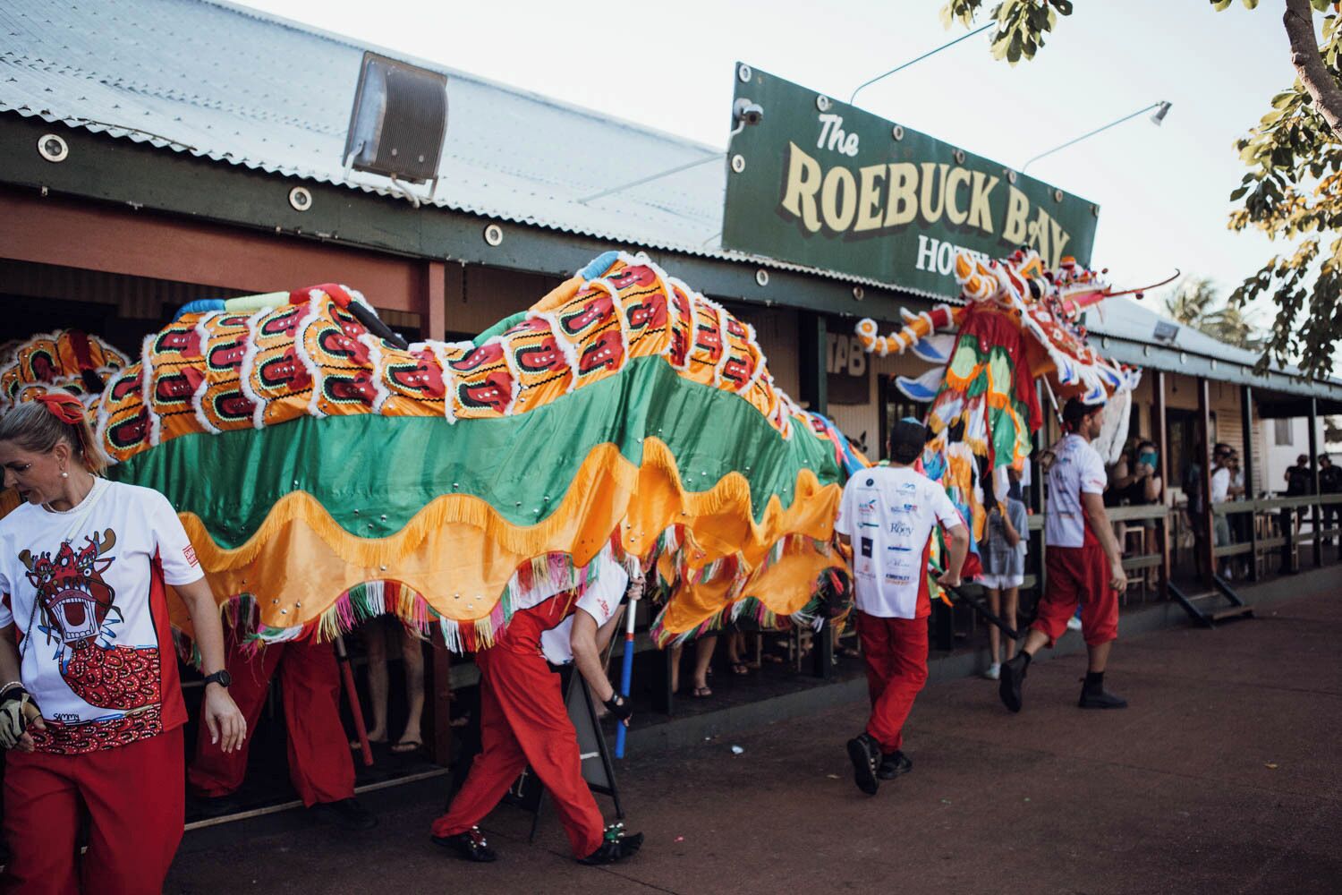 A group of people carrying a colorful dragon costume, walking past a building with a sign that reads 'The Roebuck Bay Hotel.'