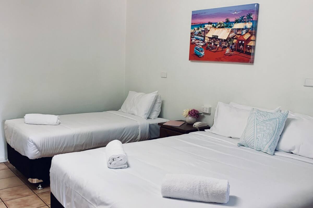 A hotel room with two beds, white linens, a painting of a market scene, and a tiled floor.