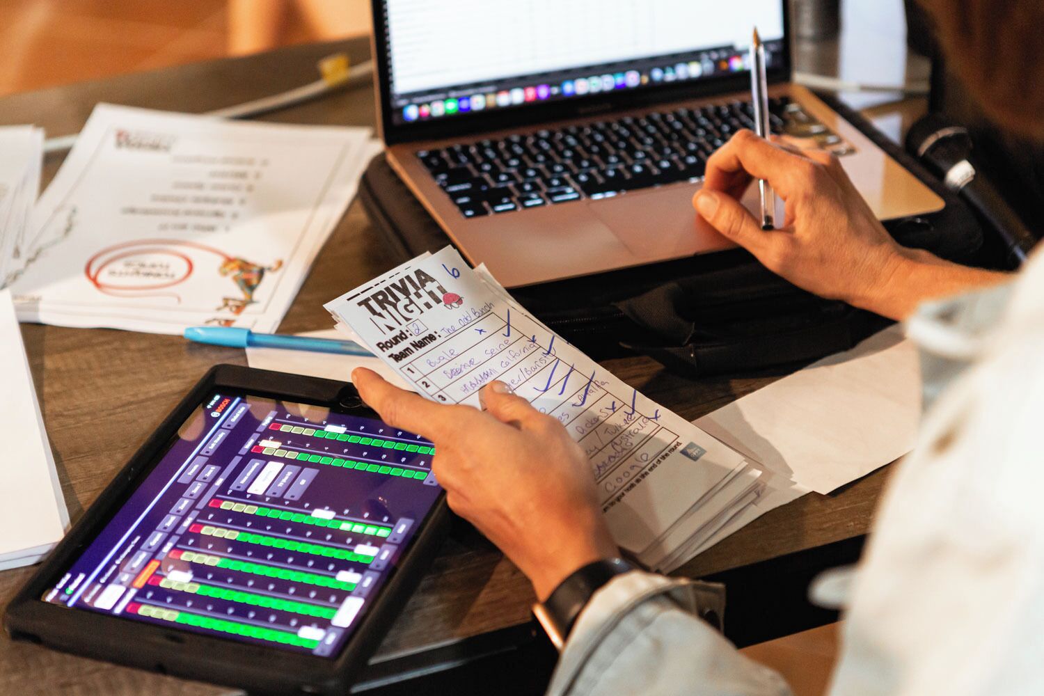 Eine Person hält ein Quiz-Blatt in der Hand, auf einem Schreibtisch stehen ein Laptop und ein Tablet, die Audio-Mixing-Software zeigen.