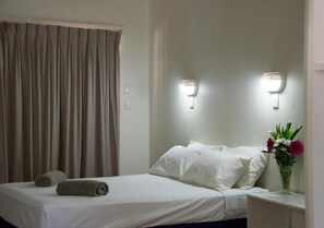A hotel room with a bed, two wall-mounted lights, a nightstand with a vase of flowers, and a curtain. - Roebuck Bay Hotel (Broome)