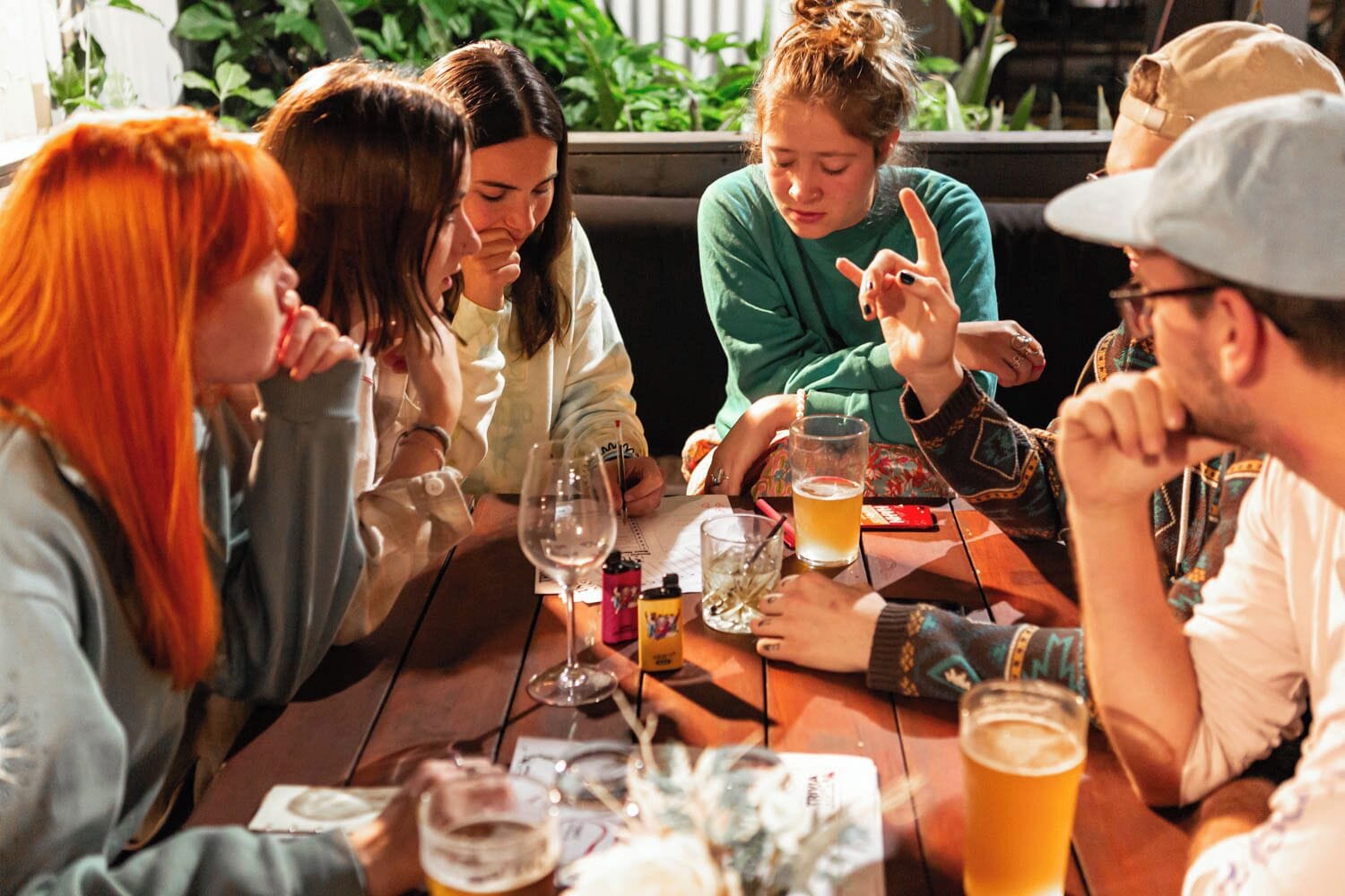 A group of people seated around a wooden table with drinks and a menu, in a setting with plants in the background.