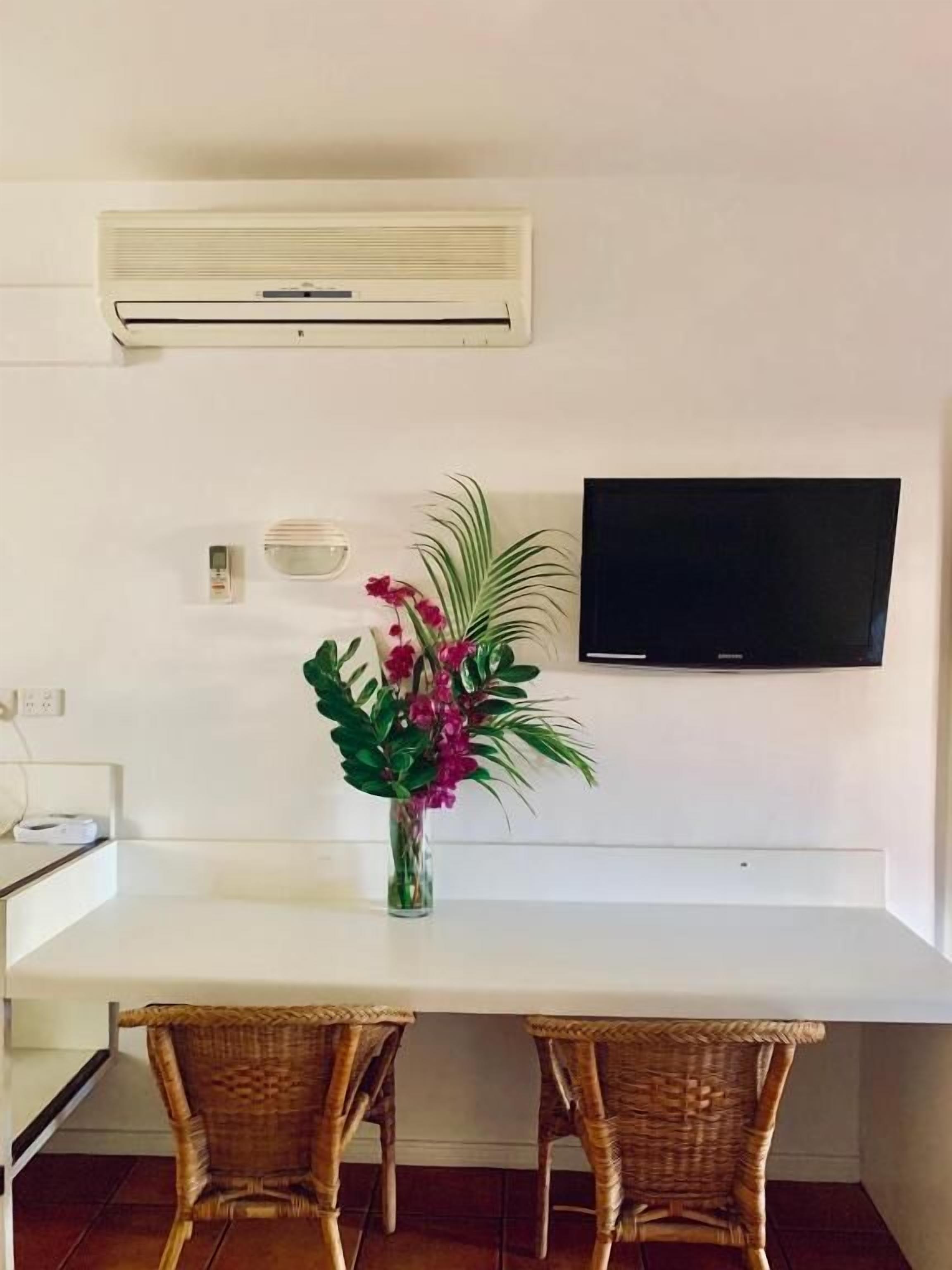 Un comedor con una mesa blanca, sillas de mimbre, un florero con flores, un aire acondicionado de pared y un televisor de pantalla plana.