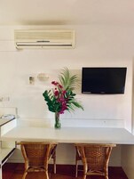 Un comedor con una mesa blanca, sillas de mimbre, un florero con flores, un aire acondicionado de pared y un televisor de pantalla plana.