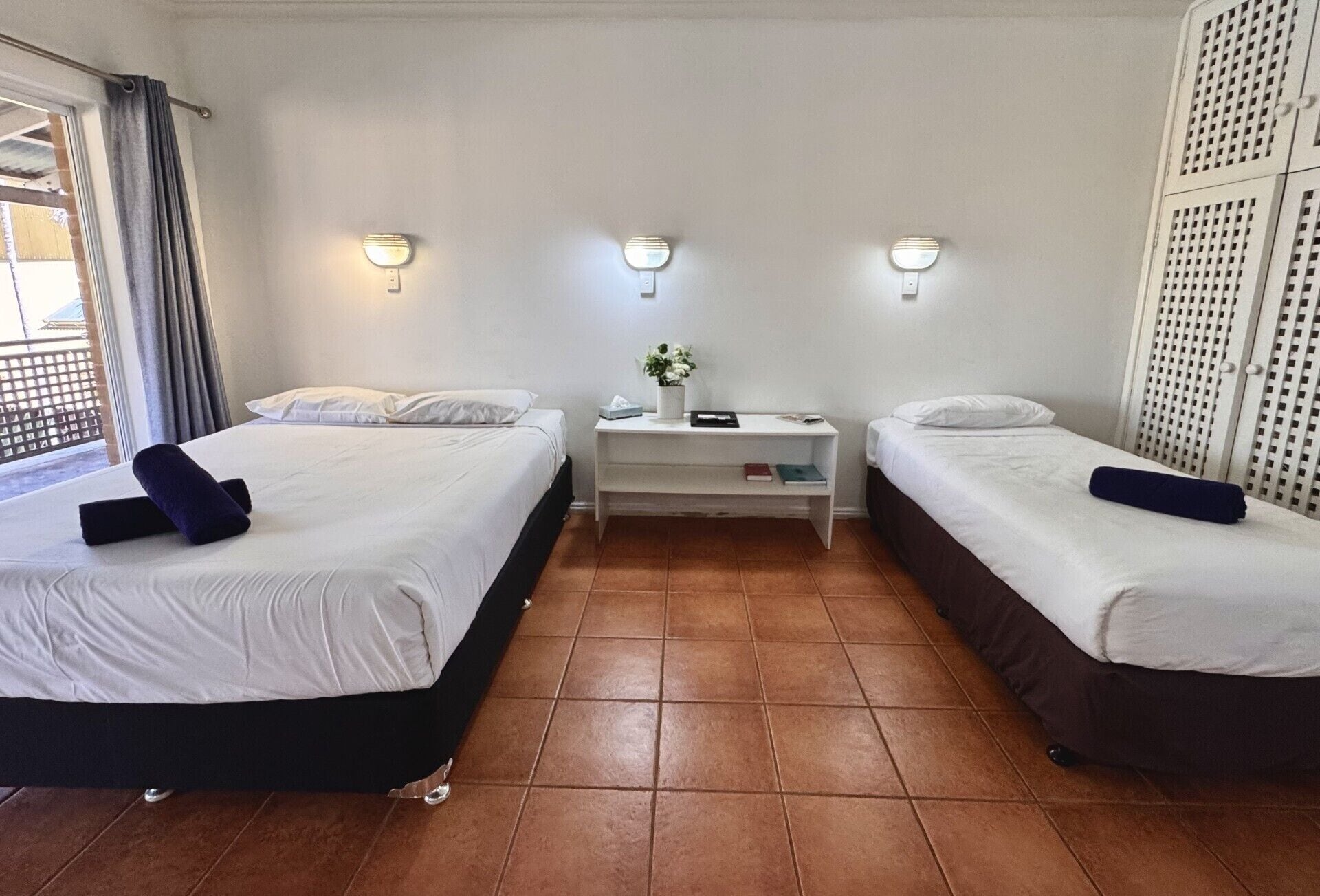 Ein Hotelzimmer mit zwei Betten, einem kleinen Nachttisch, einer Wandleuchte und Blick ins Freie.