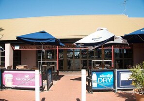 An outdoor seating area with umbrellas, a sign for Carlton Dry, and a building entrance. - Roebuck Bay Hotel (Broome)