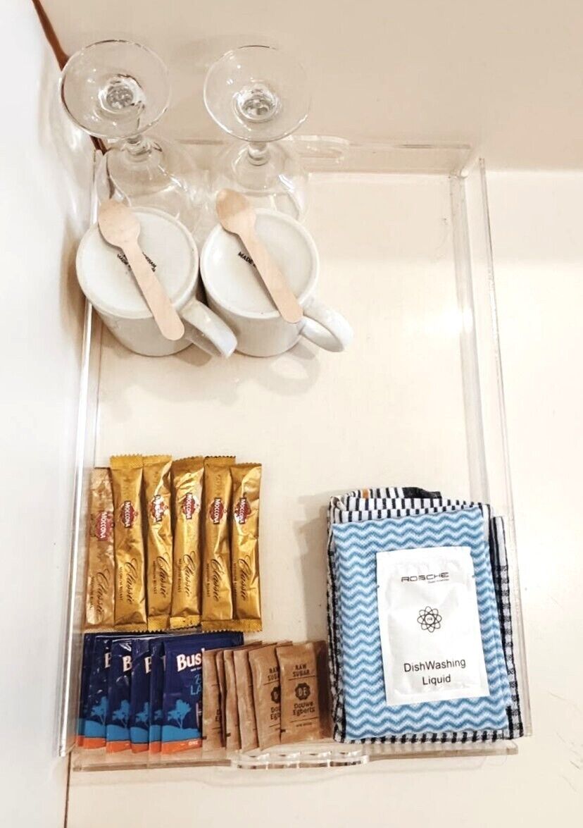A clear plastic tray with a set of white cups and spoons, a dishwashing liquid pouch, and several packets of snacks.