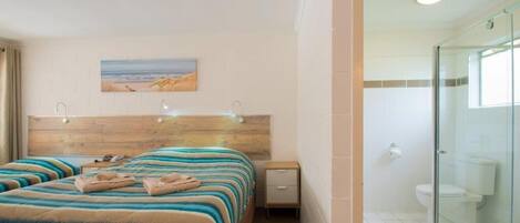 A hotel room with a bed, a nightstand, a painting of a beach, and a glass-enclosed shower.
