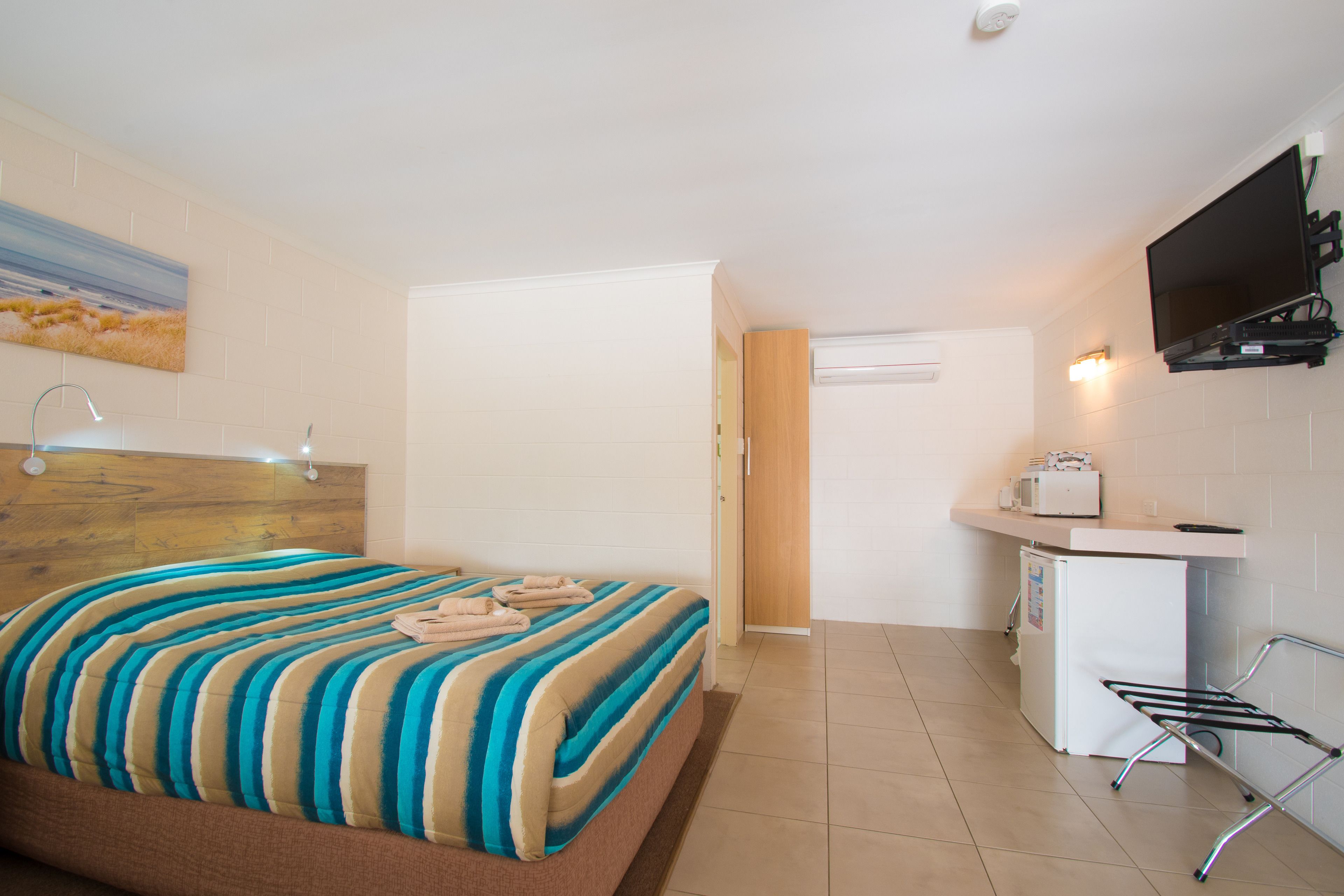 A hotel room with a bed, a small kitchenette, a TV mounted on the wall, and a door leading to another room.
