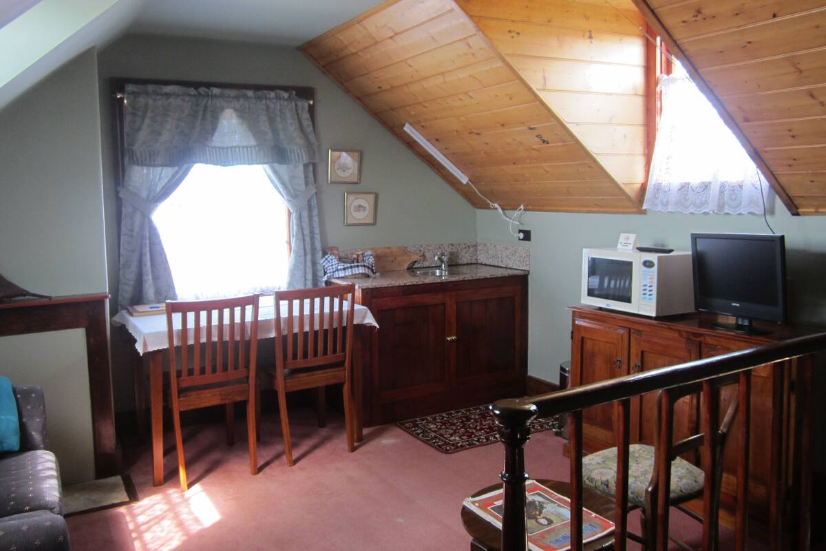 A cozy attic bedroom with a sloped ceiling, a wooden desk, a television, and a dining area with chairs.