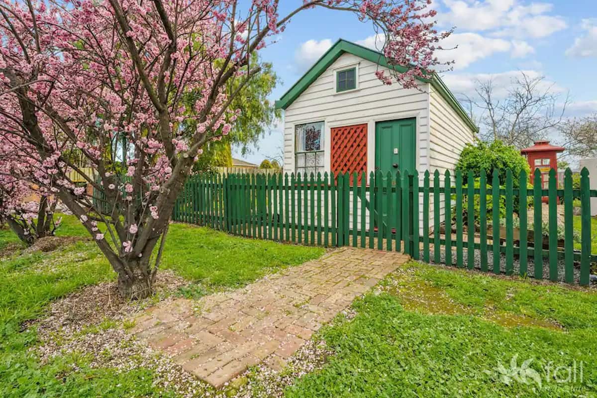 A white building with a green roof and a red door, surrounded by a green fence and blooming trees.