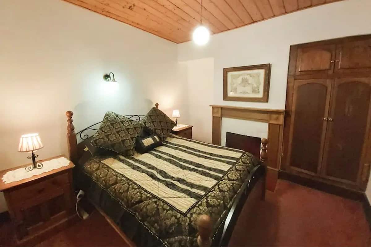 A bedroom with a wooden ceiling, a bed with a quilt, two bedside lamps, a framed picture on the wall, and a wooden wardrobe.
