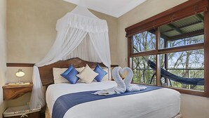 Un dormitorio con cama con dosel, mesita de noche con lámpara, cesta de mimbre con la etiqueta 'CUSHION BO', y vista a árboles por una ventana.