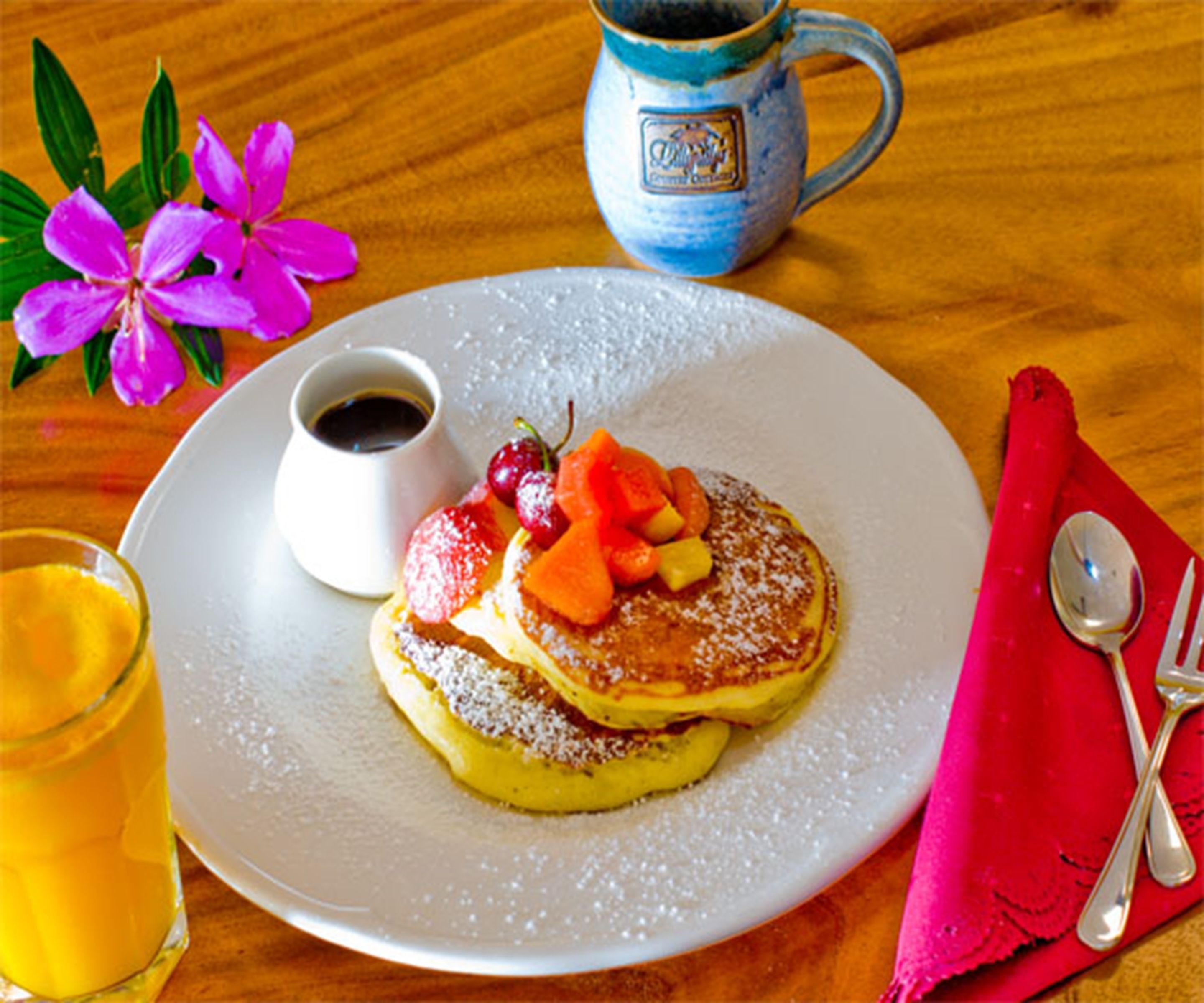 Una fuente de tortitas con fruta y almíbar, una taza de café y un vaso de zumo de naranja sobre una mesa de madera.