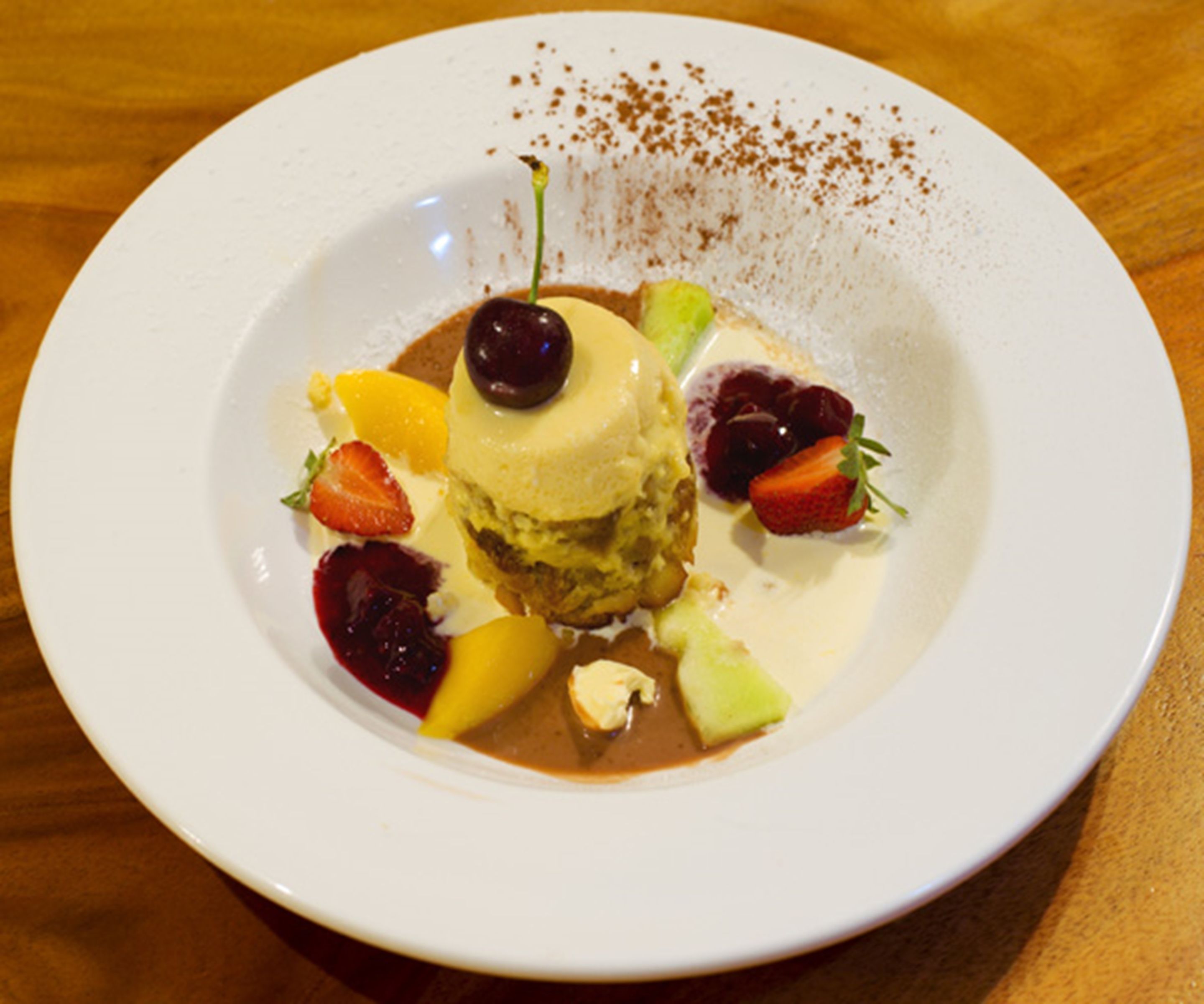 En dessert på en hvid tallerken med forskelligt frugt og et kirsebær ovenpå.