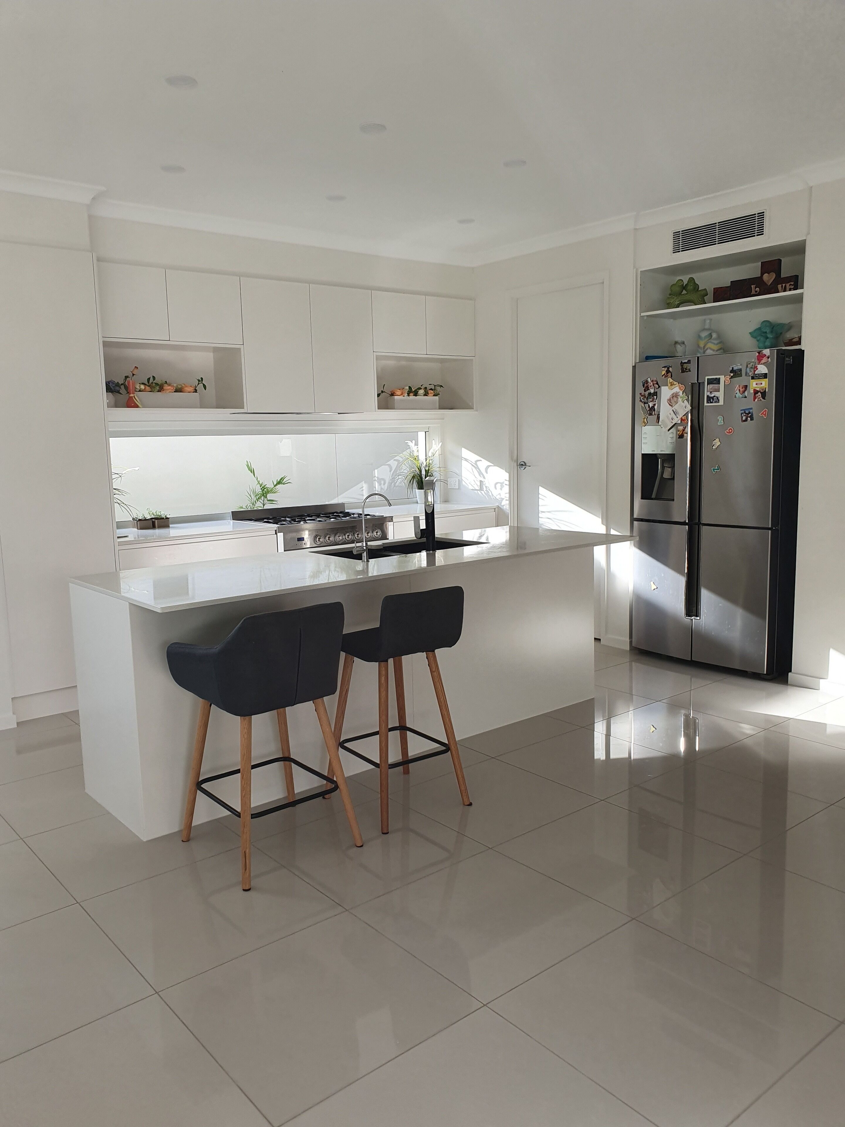 A modern kitchen with a central island, two bar stools, a stainless steel refrigerator, and white cabinetry.