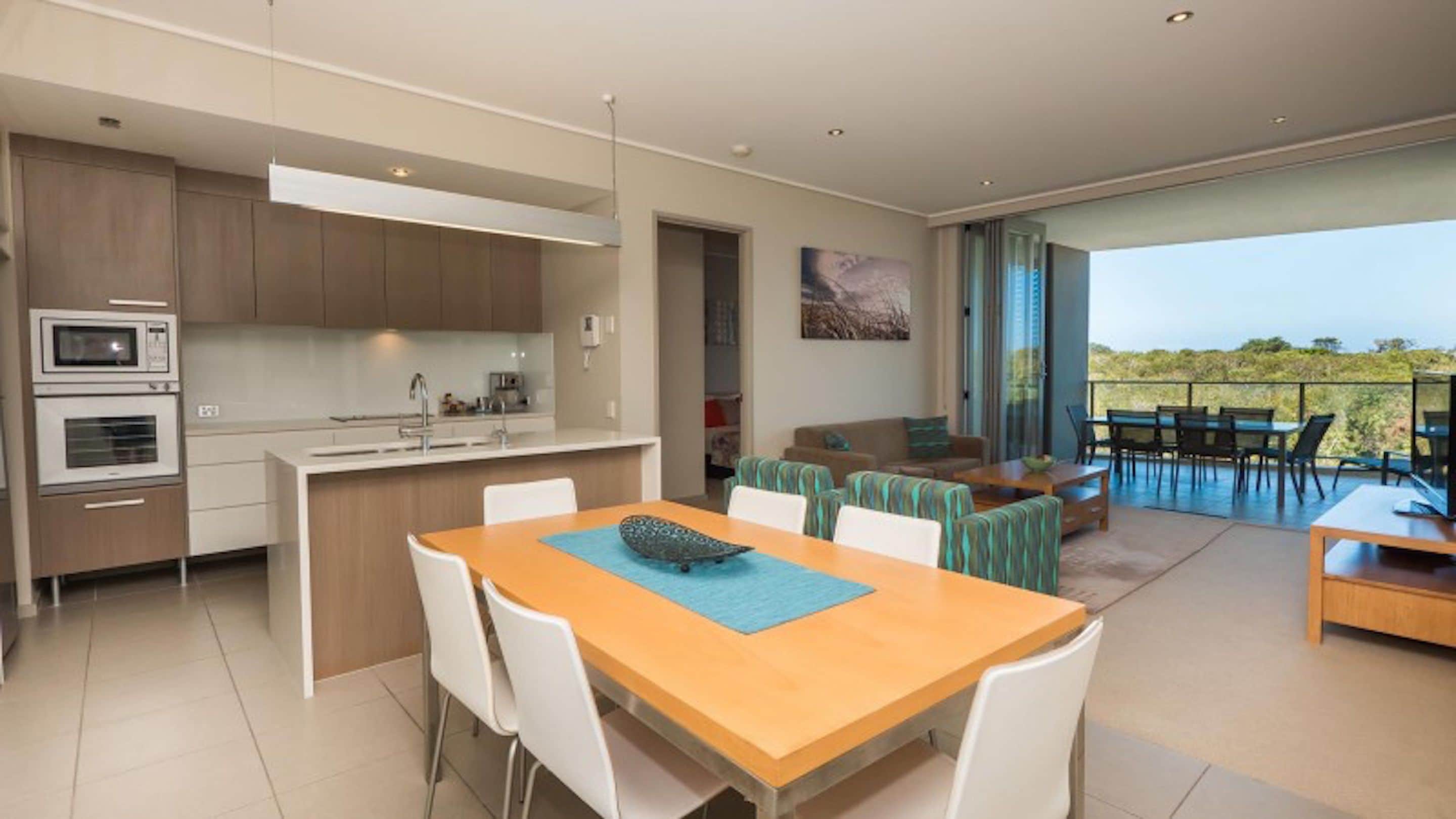 A modern kitchen with built-in appliances, a dining area with a wooden table and white chairs, and a balcony with a view of the landscape.