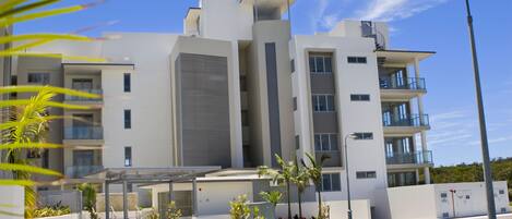 A modern multi-story building with balconies, a covered outdoor area, and a well-maintained lawn.