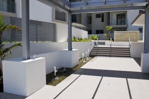 Un espacio exterior moderno con fuente de agua, escalones que llevan a un balcón y fachada de edificio blanca.