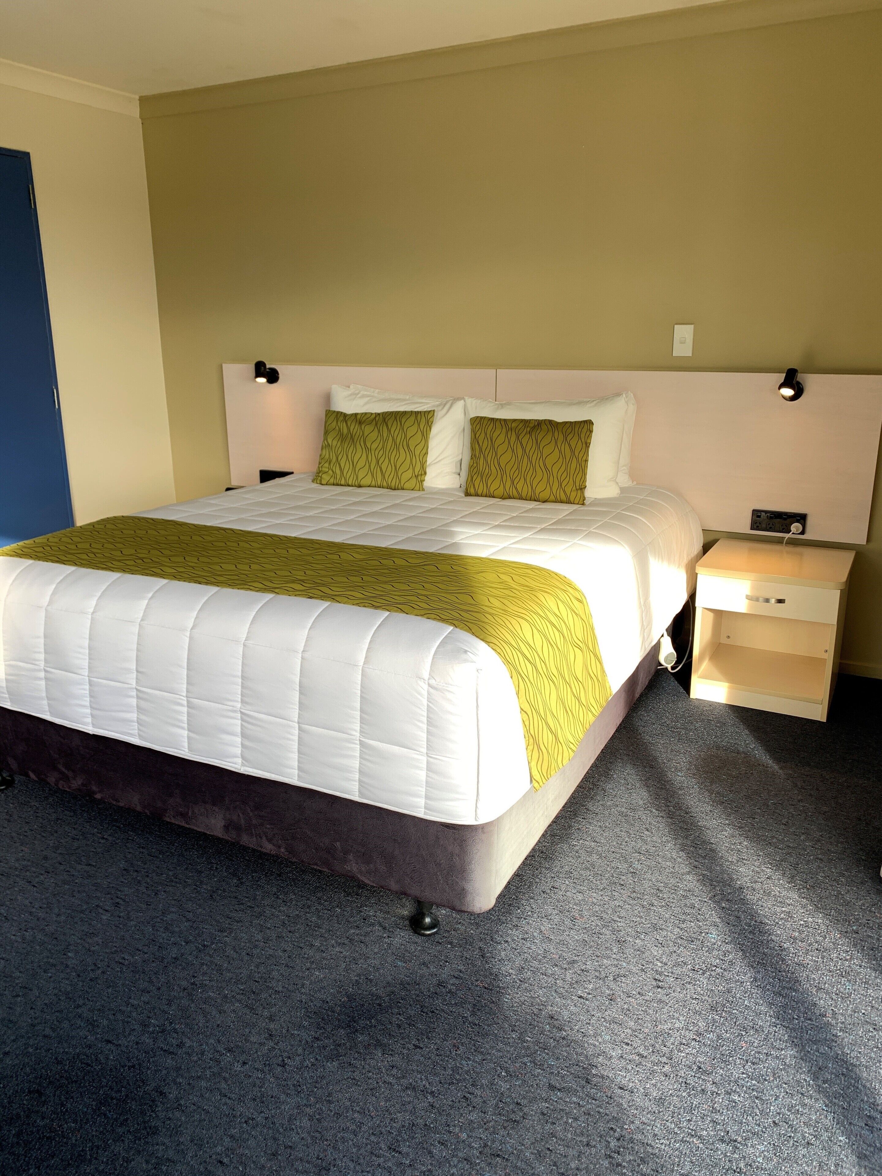 A neatly made bed with a white and yellow bedspread, a headboard with wall-mounted lights, and a small bedside table with a lamp.
