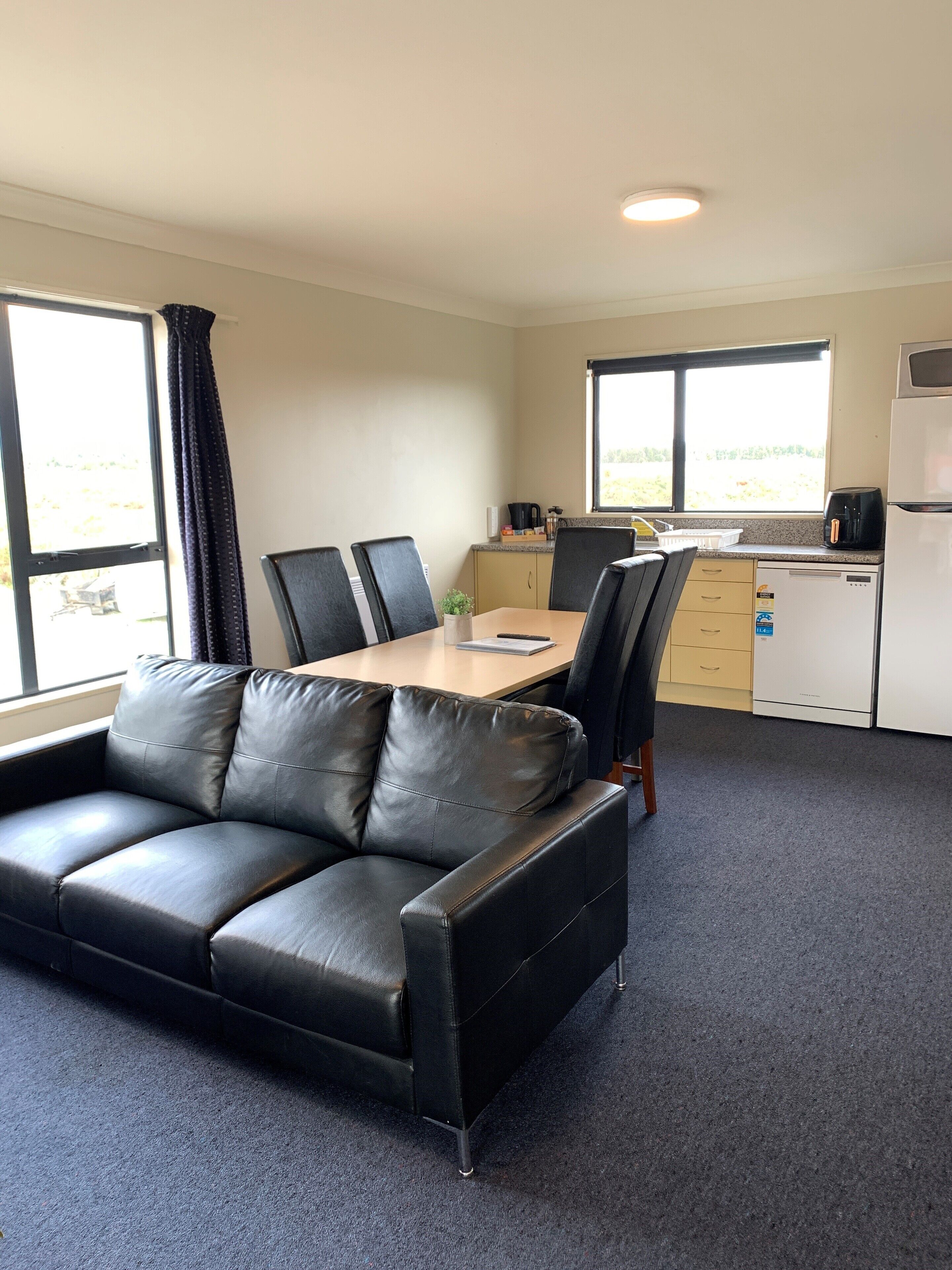 A room with a black leather sofa, a wooden dining table, and a kitchen area with a refrigerator and a coffee maker.