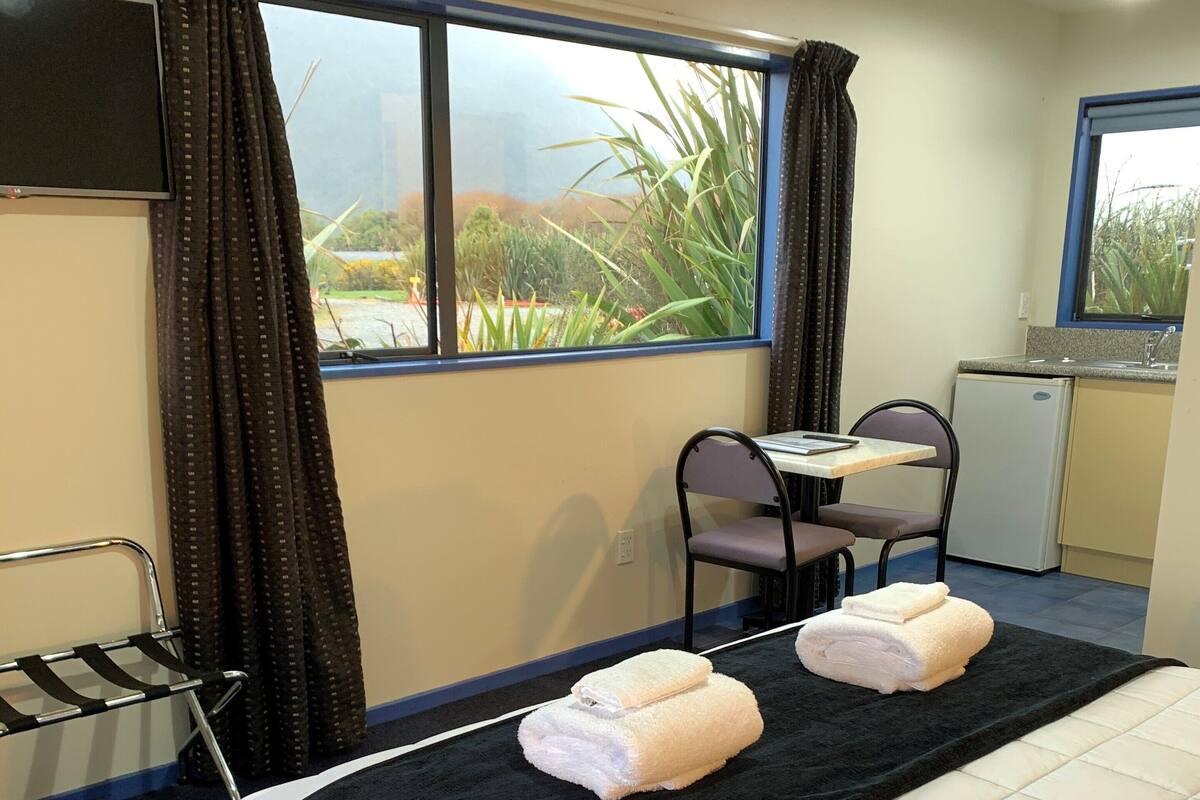 A hotel room with a bed, a desk with a chair, a small kitchenette, and a view of greenery outside.