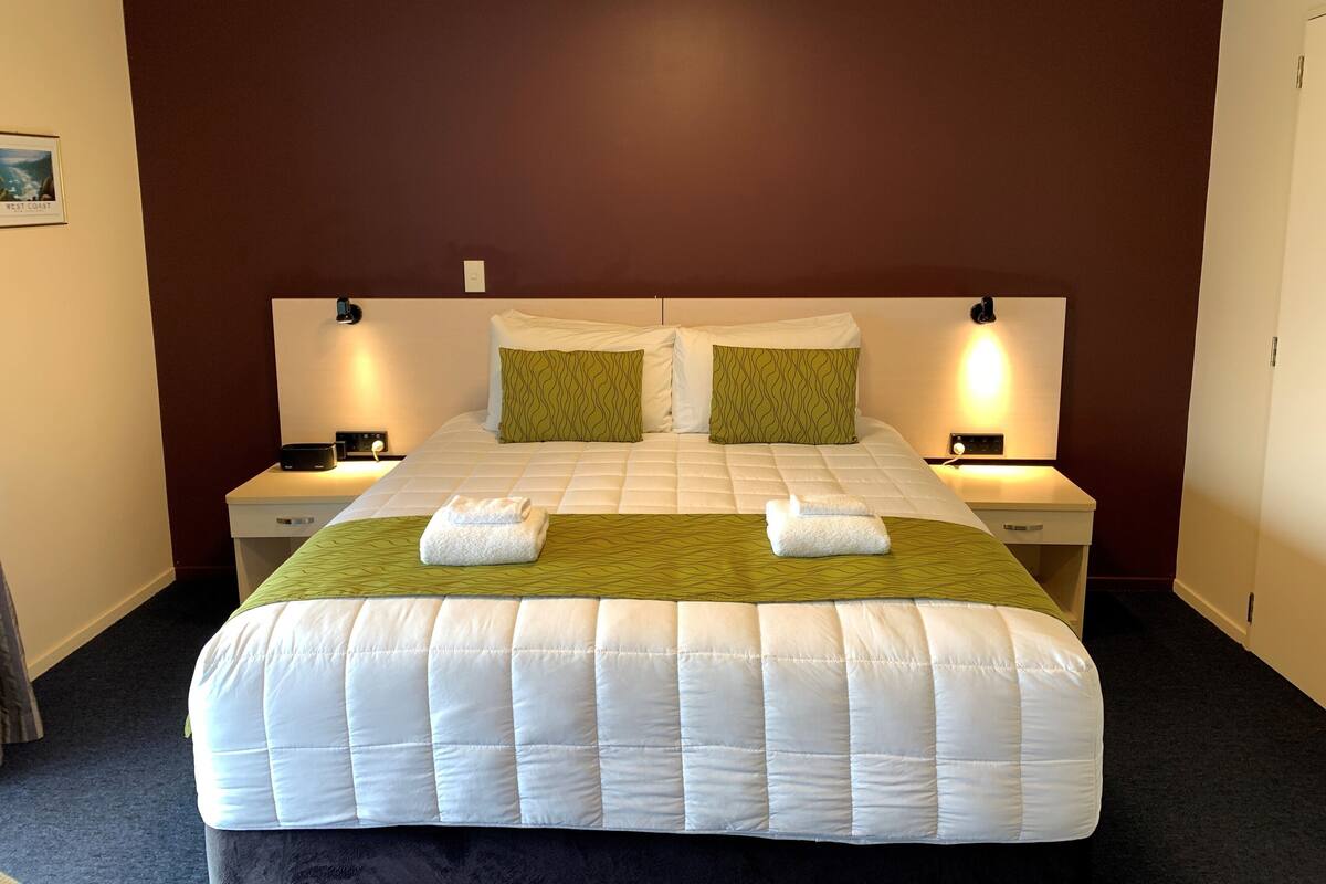 A neatly made bed with a white quilt and green accent pillows, two bedside tables with lamps, and a dark carpeted floor.