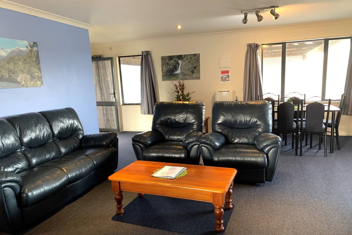 A living room with a black leather sofa, two leather armchairs, a wooden coffee table, a dining table with chairs, and a large window with curtains.