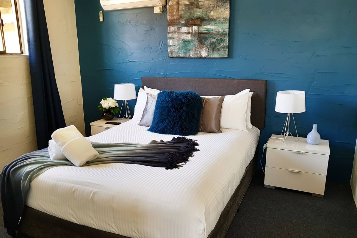 A neatly made bed with white linens, a blue throw pillow, and a dark blanket. There are two bedside tables with lamps and a vase on one. The room has a blue wall with a textured painting above the bed.