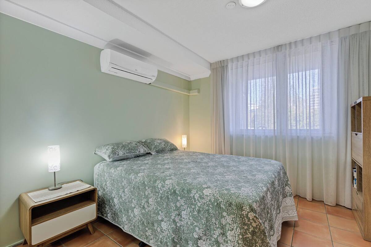 A bedroom with a bed, an air conditioner, a bedside lamp, a nightstand, and a window with curtains.