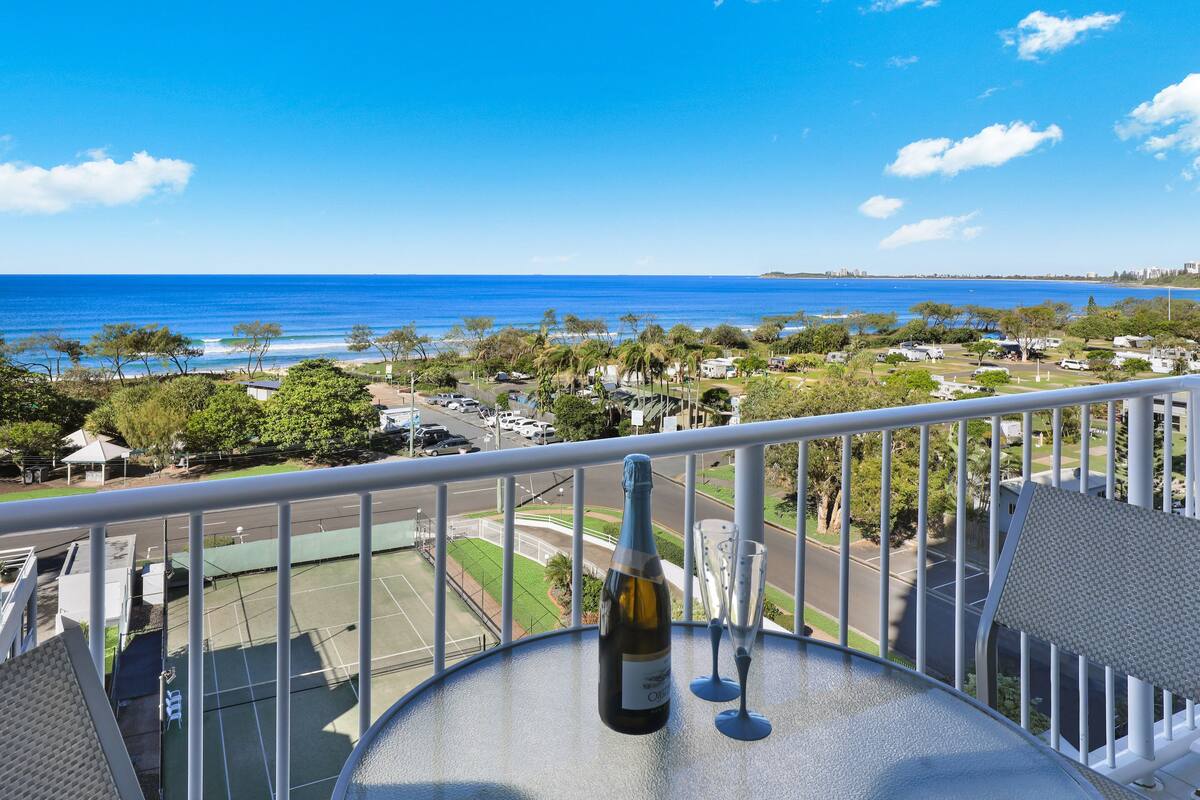 A balcony with a table, a bottle of wine, and two glasses. The view includes a beach, ocean, and a parking area with cars and greenery.