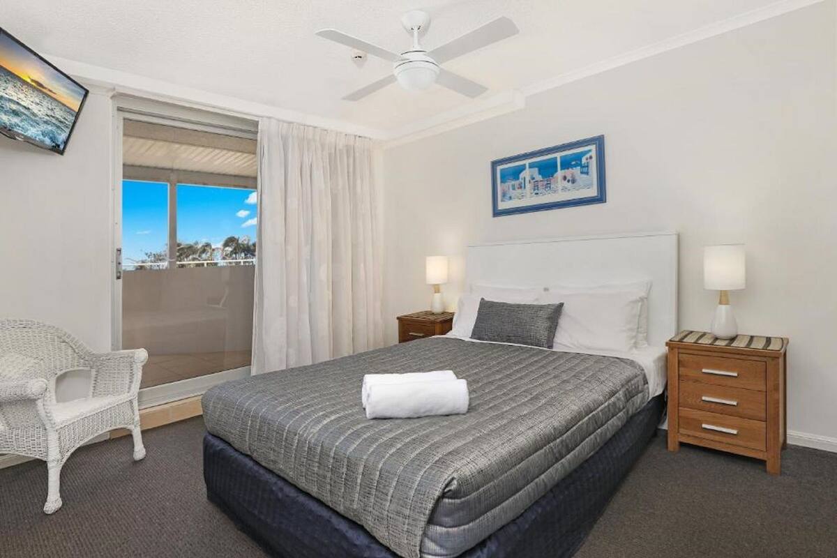A hotel room with a bed, two bedside tables, a chair, a ceiling fan, and a view of the outdoors.
