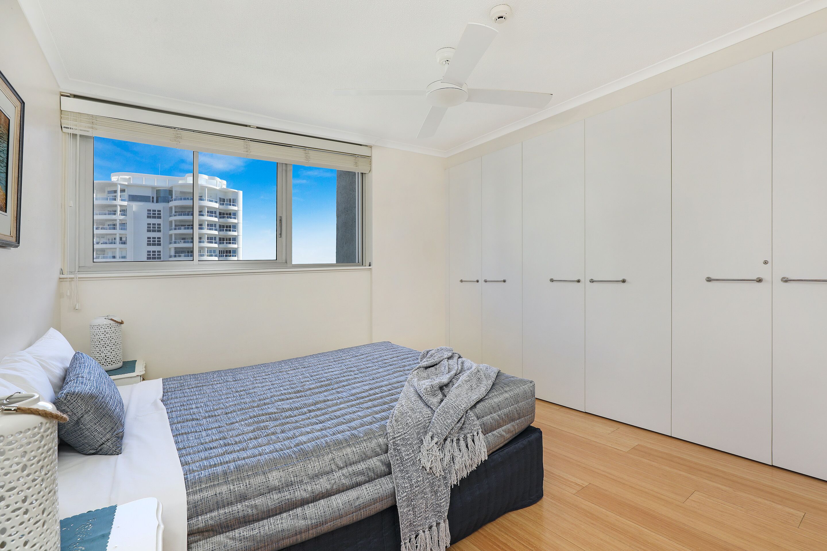 A bedroom with a bed, a window with a view of a building, a ceiling fan, and a wardrobe.