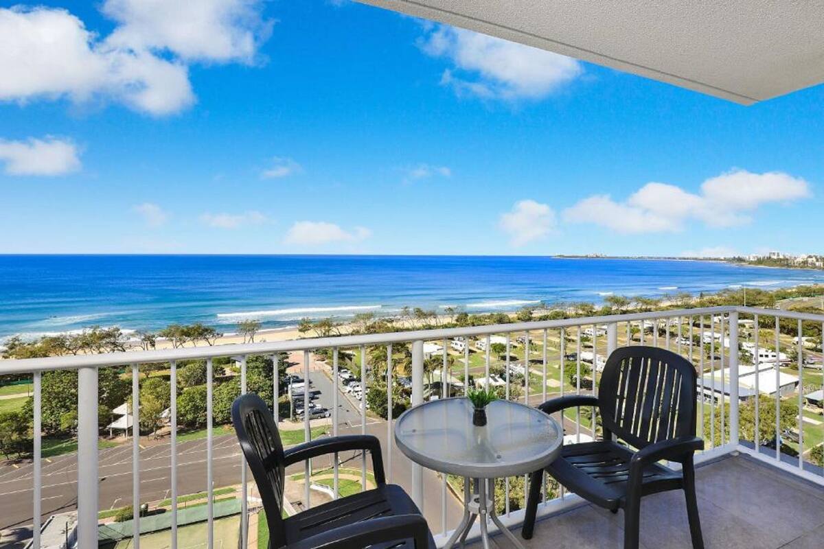 A balcony with two chairs and a small table, overlooking a beach and ocean.