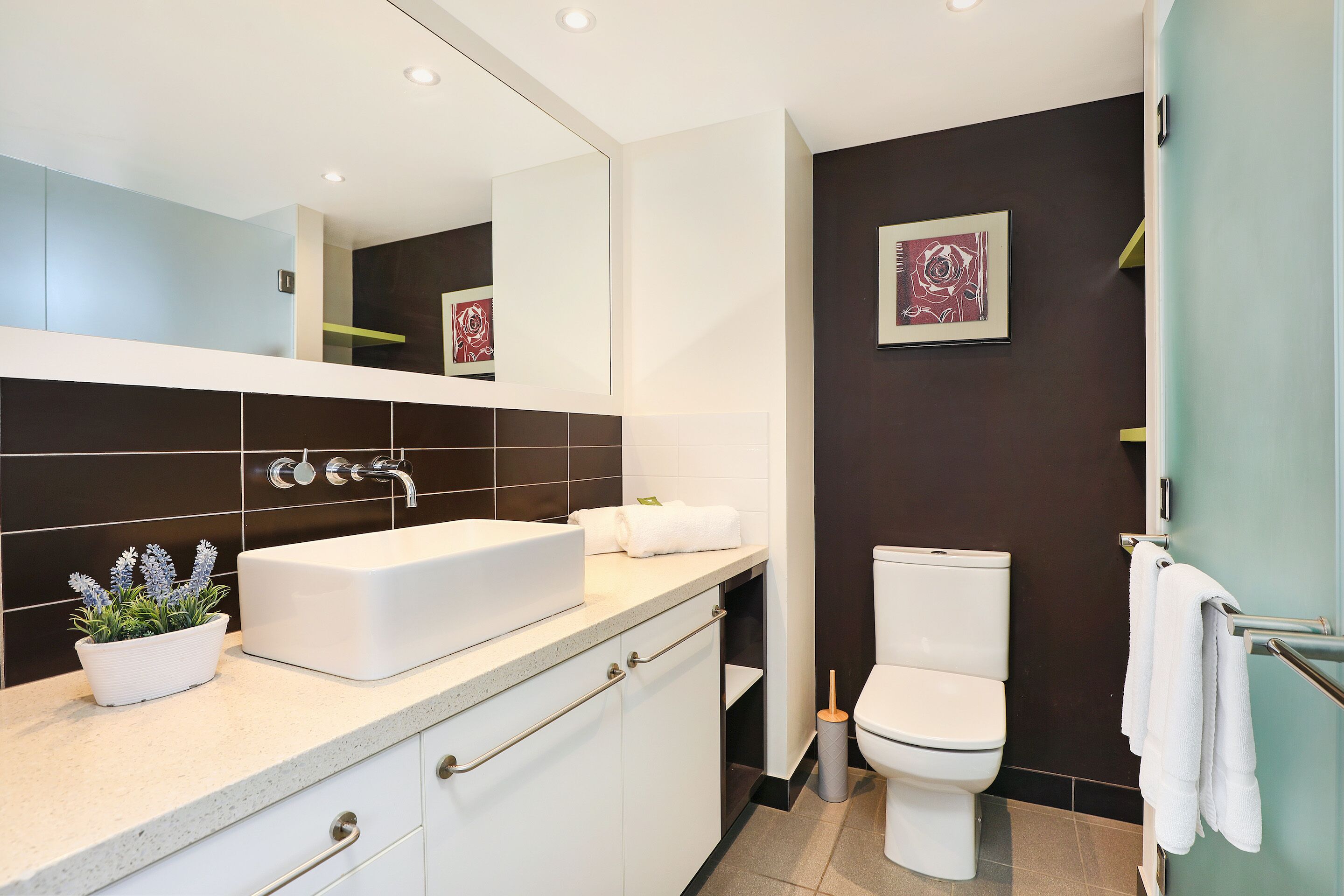 A modern bathroom with a white sink, dark tiled walls, a toilet, and a mirror.