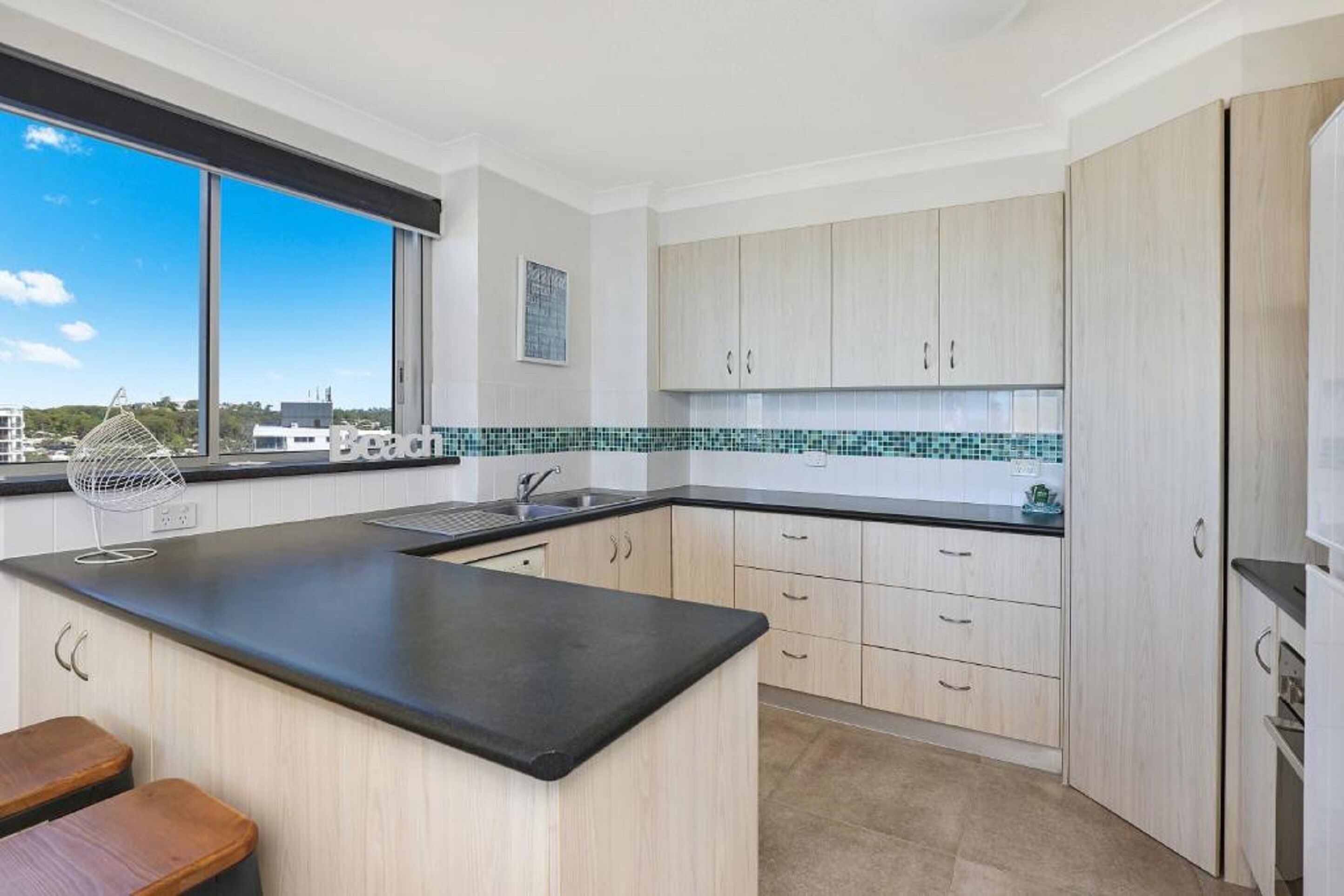 A modern kitchen with a black countertop, light wood cabinets, a tiled backsplash, and a window overlooking a cityscape.